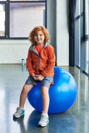 Foto de Tiro vertical de pelo rojo niño alegre sentado en la pelota de fitness y sonriendo alegremente a la cámara - Imagen libre de derechos