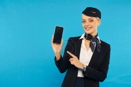 elegante azafata en elegante uniforme apuntando al teléfono inteligente con pantalla en blanco en el telón de fondo cian