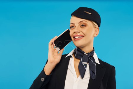 junge und gut gelaunte Stewardess in eleganter Uniform spricht auf Smartphone mit leerem Bildschirm auf blauem Hintergrund
