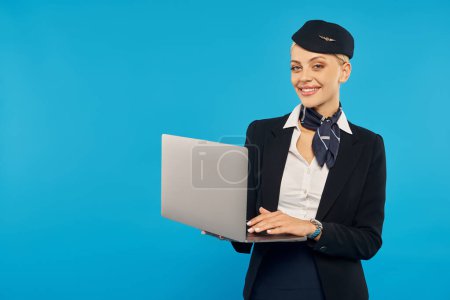 azafata alegre en elegante uniforme corporativo celebración de la computadora portátil y mirando a la cámara en el fondo azul