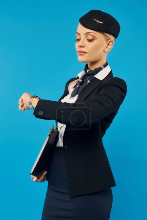 Photo for Young stewardess in elegant uniform looking at wristwatch while checking time on blue backdrop - Royalty Free Image