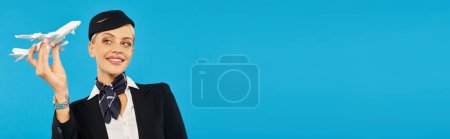 Photo for Happy and charming air hostess in stylish uniform holding airplane model on blue backdrop, banner - Royalty Free Image