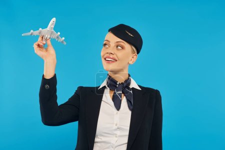 junge inspirierte Frau in eleganter Stewardess-Uniform mit Flugzeugmodell vor blauem Hintergrund
