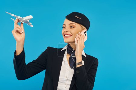 Glückliche Stewardess in Uniform hält Flugzeugmodell in der Hand und spricht mit Smartphone auf blauem Hintergrund