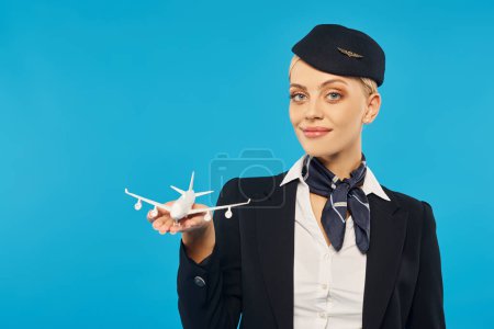 elegant stewardess in uniform holding airplane model and smiling at camera on cyan backdrop