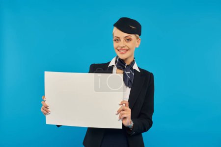 happy stewardess in stylish unform holding blank placard and looking at camera on blue backdrop