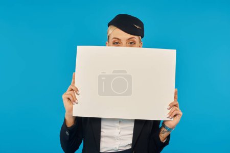 junge Frau in Uniform der Stewardess verdeckt Gesicht mit leerem Plakat auf blauem Hintergrund