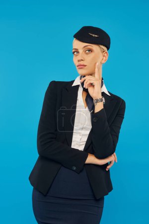Photo for Young and dreamy woman in uniform of air hostess touching face and looking away on blue backdrop - Royalty Free Image