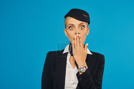 shocked stewardess with bugling eyes covering mouth with hand and looking at camera on blue