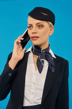 serious stewardess in elegant uniform talking on mobile phone on blue, work in airline industry