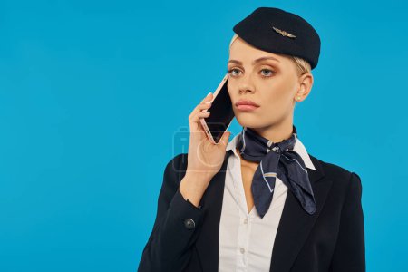 young upset stewardess in stylish uniform talking on smartphone on blue studio background
