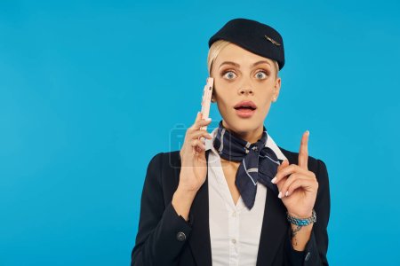 verblüffte Stewardess in Uniform spricht auf Handy und zeigt Ideengeste auf blauem Hintergrund