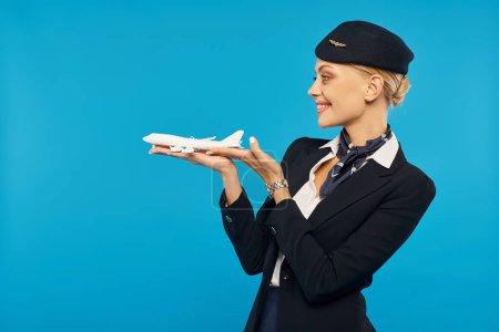 Photo for Cheerful attractive stewardess in uniform standing with airplane model on blue backdrop, side view - Royalty Free Image