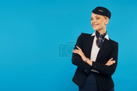 Photo for Joyful air hostess in corporate uniform posing with folded arms on blue backdrop, airline industry - Royalty Free Image