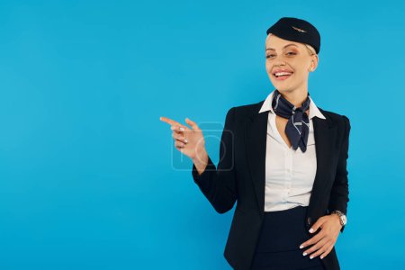 smiling stewardess pointing away while showing direction and looking at camera on blue background