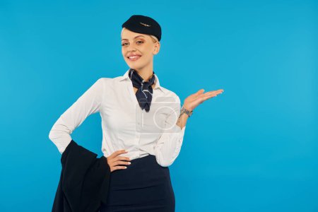 Photo for Happy air hostess in white blouse and stewardess hat pointing with hand and holding jacket on blue - Royalty Free Image