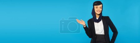 Photo for Elegant positive stewardess of arabian airlines pointing with hand and showing way on blue, banner - Royalty Free Image