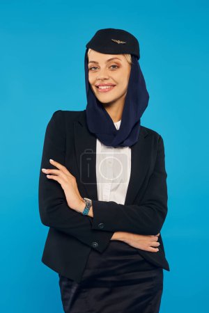 encantadora azafata de las aerolíneas árabes en uniforme con pañuelo en la cabeza posando con los brazos cruzados en azul