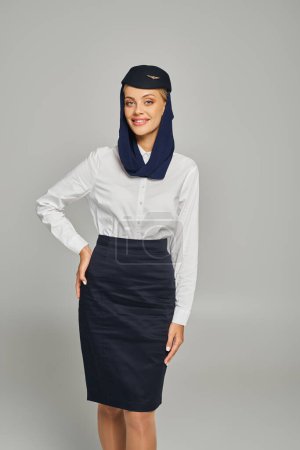 stylische und gut gelaunte Stewardess in Uniform arabischer Airlines posiert mit der Hand an der Hüfte auf grau