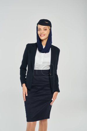 smiling air hostess in headscarf and corporate uniform of arabian airlines looking at camera on grey