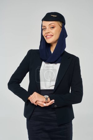 graziöse Stewardess arabischer Fluggesellschaften mit glücklichem Lächeln und Kopftuch, die in die Kamera auf grau blickt