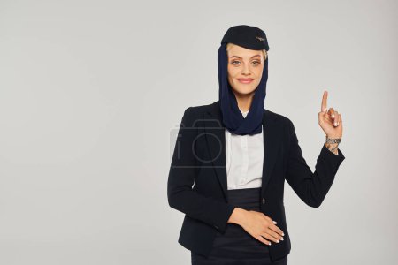 fröhliche Stewardess in der Kleiderordnung arabischer Fluggesellschaften, die mit dem Finger auf grauen Hintergrund zeigt