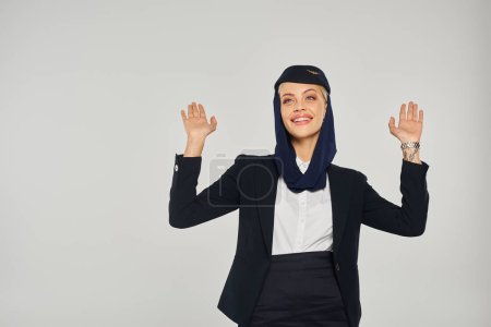 excited stewardess in uniform of arabian airlines looking up and waving hands on grey backdrop