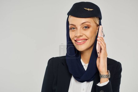 charming stewardess in arabian airlines uniform talking on smartphone and smiling at camera on grey