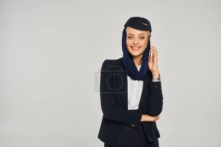 Photo for Graceful air hostess of arabian airlines talking on mobile phone and smiling at camera on grey - Royalty Free Image