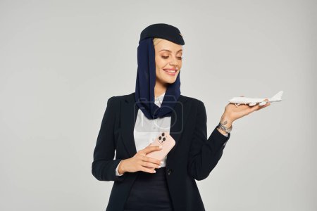 smiling arabian airlines stewardess with mobile phone looking at airplane model on grey backdrop