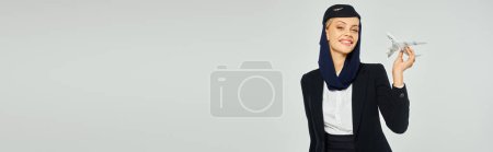 Téléchargez les photos : Hôtesse de l'air joyeuse des compagnies aériennes arabes en uniforme élégant debout avec modèle d'avion sur gris, bannière - en image libre de droit