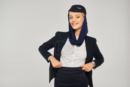 Téléchargez les photos : Hôtesse de l'air souriante des compagnies aériennes arabes en uniforme et foulard posant les mains sur la taille sur gris - en image libre de droit