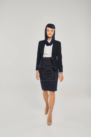 full length of graceful arabian airlines stewardess in uniform walking and looking at camera on grey
