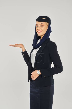 Téléchargez les photos : Hôtesse attrayante dans les compagnies aériennes arabes uniforme souriant à la caméra et pointant avec la main sur le gris - en image libre de droit