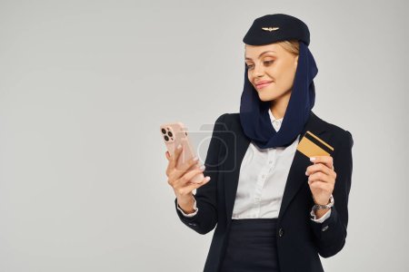 smiling stewardess in arabian airlines uniform holding credit card and looking at smartphone on grey