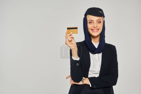 young and elegant arabian airlines stewardess with credit card smiling at camera on grey backdrop