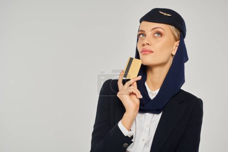 Téléchargez les photos : Hôtesse de l'air arabe réfléchie des compagnies aériennes en uniforme tenant la carte de crédit et détournant les yeux sur gris - en image libre de droit