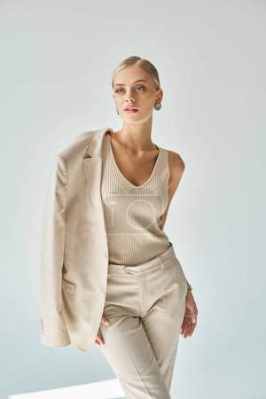 attraktive Frau in beiger Freizeitkleidung mit Blazer auf der Schulter schaut vor grauem Hintergrund weg