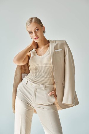 junges Mode-Model in stilvoller beiger Kleidung, die den Hals berührt und mit der Hand in der Tasche auf grau posiert