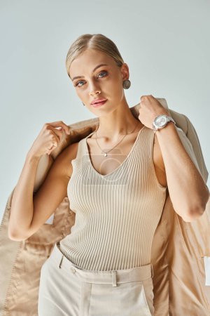 stylisches blondes weibliches Modell in pastellbeiger Kleidung mit Blazer und Blick in die Kamera auf grau