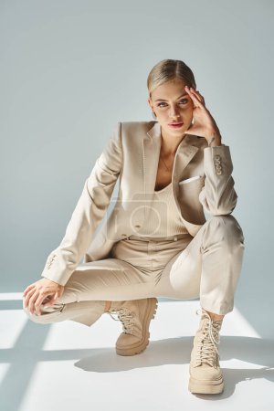 Téléchargez les photos : Femme élégante en vêtements beige et bottes lacées posant sur des hanches et regardant la caméra sur gris - en image libre de droit