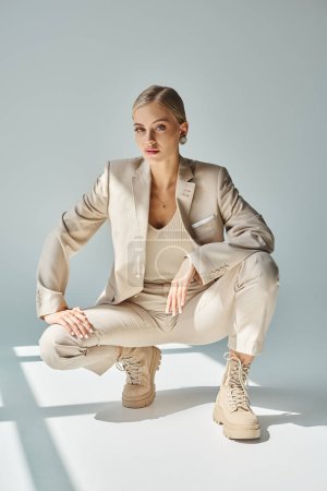 Téléchargez les photos : Modèle de mode glamour en costume beige et bottes lacées assis sur des hanches au soleil sur gris - en image libre de droit