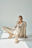 sensual woman in pastel beige wear and laced boots posing and looking at camera in sunshine on grey hoodie #677651496