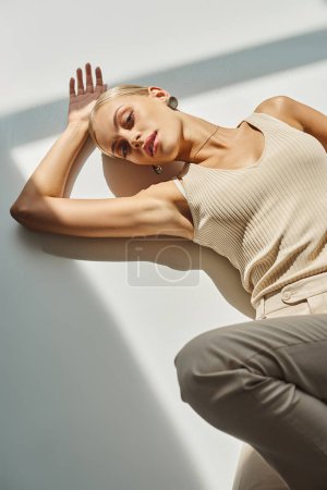 encantadora mujer rubia en camiseta beige tumbado y mirando a la cámara en el sol en gris