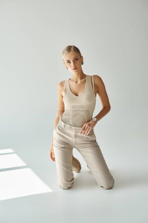 Photo for Trendy blonde woman in tank top and pants kneeling on grey backdrop with sunlight, urban fashion - Royalty Free Image