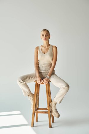 Foto de Longitud completa del modelo femenino en camiseta sin mangas beige y pantalones sentados en taburete alto sobre fondo gris - Imagen libre de derechos