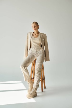 Photo for Pretty blonde woman in pastel beige suit looking at camera on tall stool in sunshine on grey - Royalty Free Image