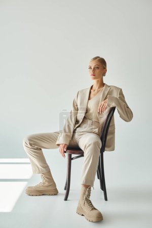Foto de Modelo de moda rubia en traje beige pastel sentado en la silla y mirando hacia otro lado en el sol en gris - Imagen libre de derechos