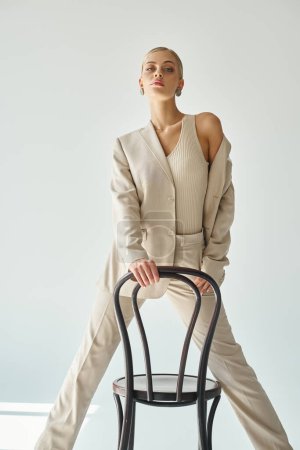 Foto de Modelo femenino atractivo en traje elegante posando con silla y mirando a la cámara en el fondo gris - Imagen libre de derechos