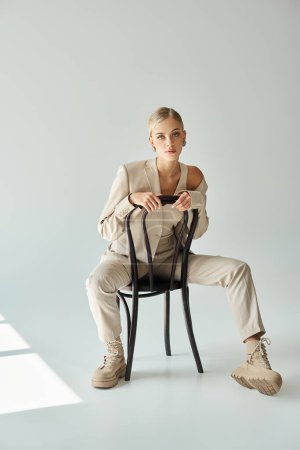young woman in total beige outfit sitting on chair and looking at camera on grey, urban style